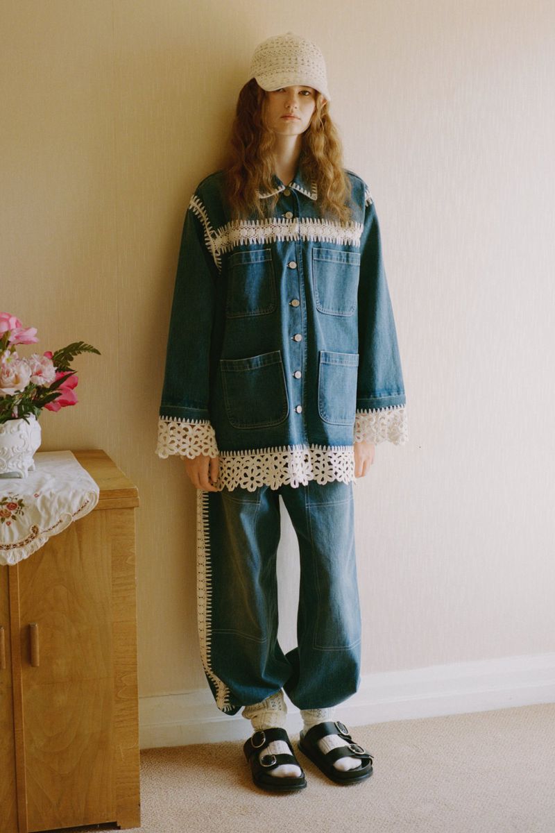 Young woman in a blue denim jacket and jeans accented with crocheted trim and a white crocheted hat, standing indoors, casually posed.