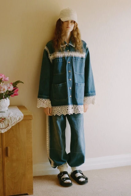 Young woman in a blue denim jacket and jeans accented with crocheted trim and a white crocheted hat, standing indoors, casually posed.