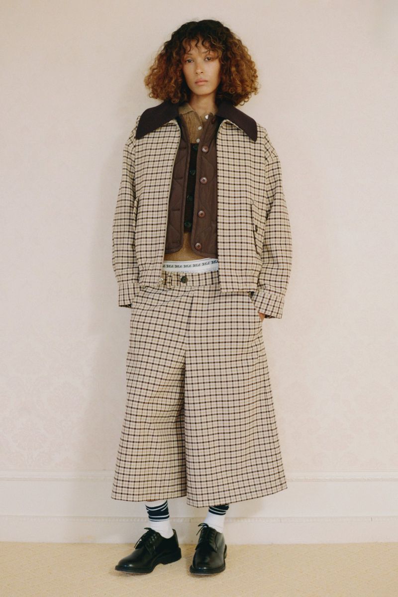 A young adult female stands indoors wearing a brown and tan plaid jacket, matching culottes, and black dress shoes in a fashion editorial shot.