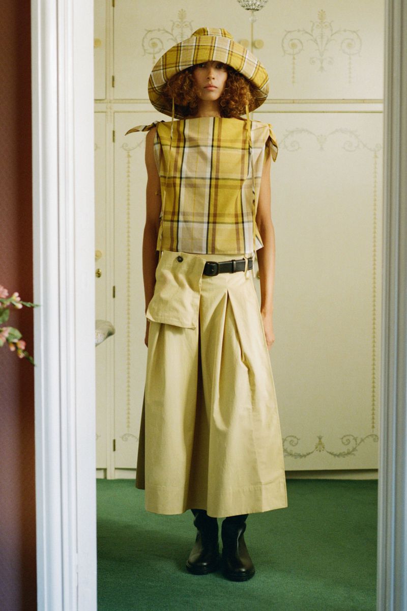 A woman in a yellow plaid top and matching hat poses in front of a white door, wearing a tan skirt and black boots. Fashion shoot.