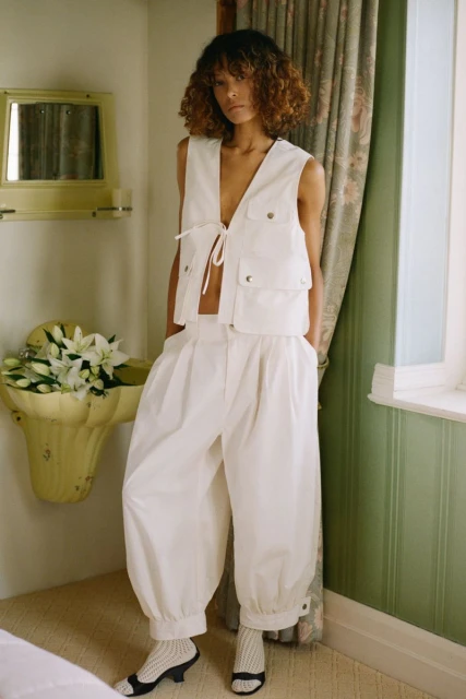 Woman in white vest and balloon pants poses in an indoor setting with natural light, floral arrangement, and patterned curtains.
