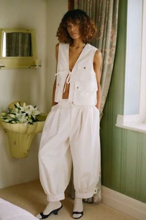 Woman in white vest and balloon pants poses in an indoor setting with natural light, floral arrangement, and patterned curtains.