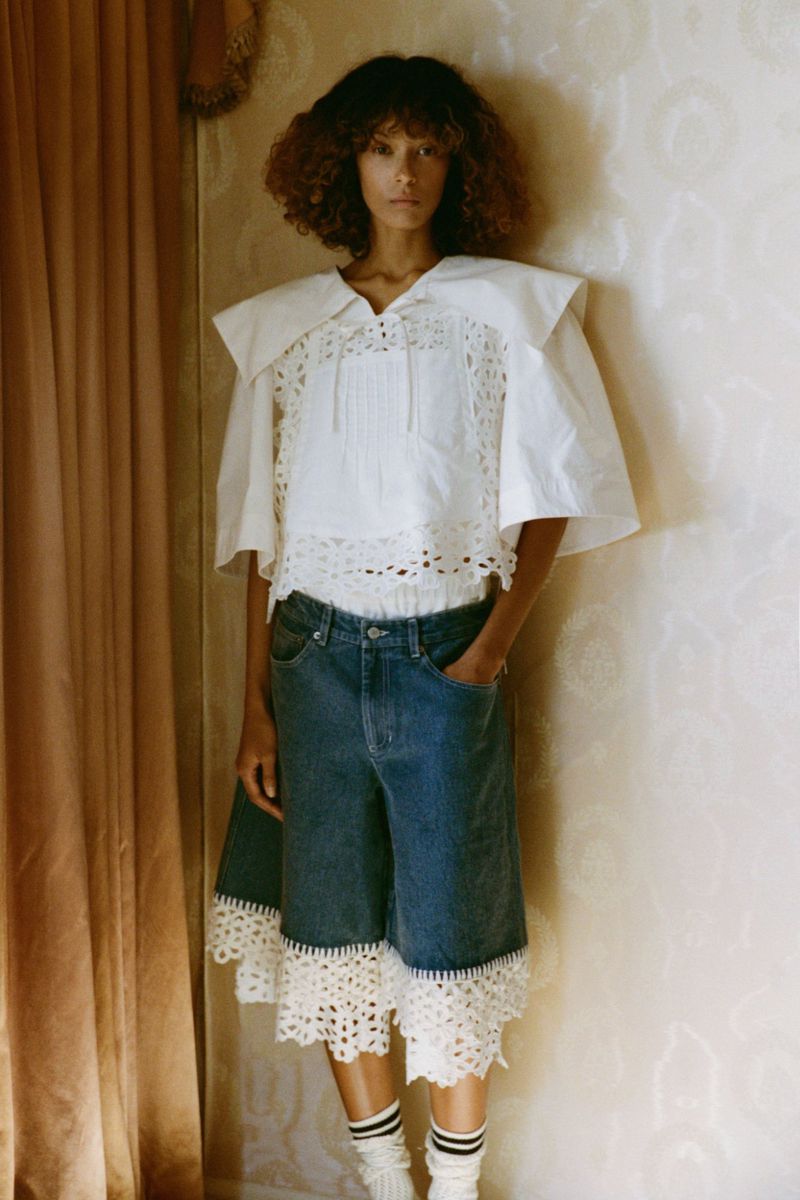 Woman poses indoors wearing a white blouse with lace, blue denim shorts with lace, and striped socks, with a neutral wall backdrop.