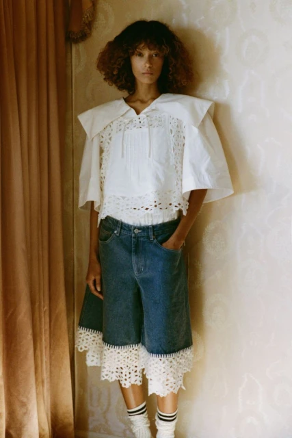 Woman poses indoors wearing a white blouse with lace, blue denim shorts with lace, and striped socks, with a neutral wall backdrop.
