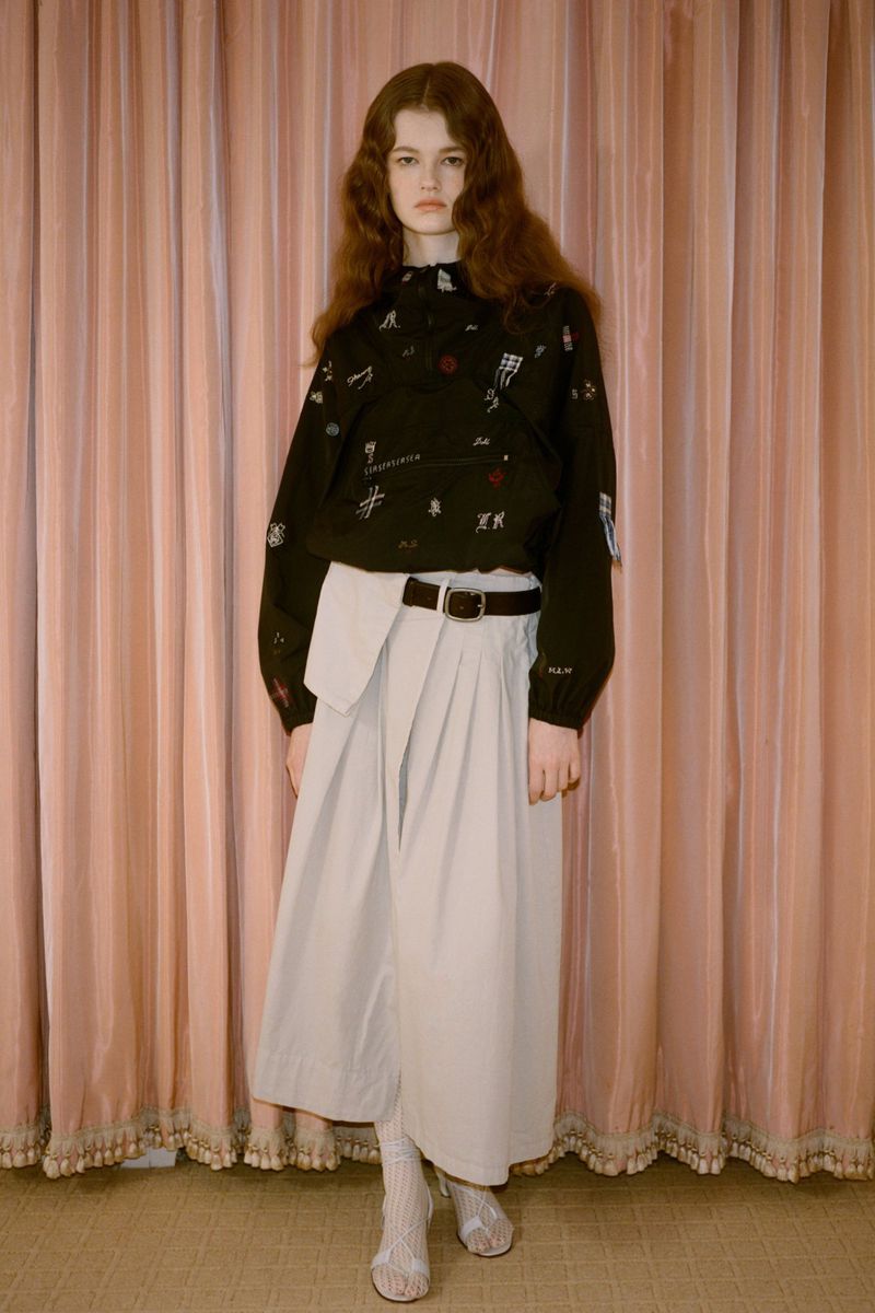 Young woman in black long-sleeve shirt with drawings, grey wrap skirt, stands in front of a peach-colored curtain in photo studio.