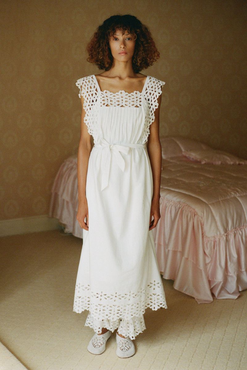 A woman stands indoors wearing a white crochet-trimmed dress and slippers, posed in front of a pink bed and patterned wall.