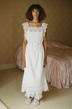 A woman stands indoors wearing a white crochet-trimmed dress and slippers, posed in front of a pink bed and patterned wall.