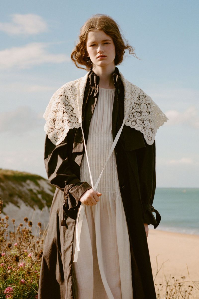 Fashion shot of a young woman in a black coat with white lace collar and a dress, posing outdoors by the sea under a bright sky.