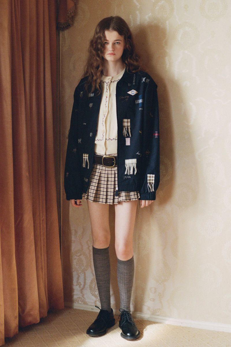 A young woman in a patterned jacket and plaid skirt stands in a warmly lit room with neutral wallpaper and a flowing curtain.