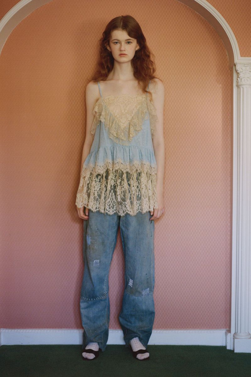 A young adult woman with long curly auburn hair poses in a studio wearing a lace-trimmed tank top and distressed denim jeans.