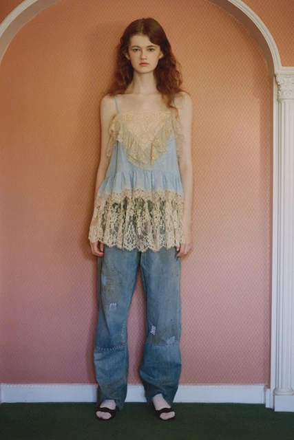 A young adult woman with long curly auburn hair poses in a studio wearing a lace-trimmed tank top and distressed denim jeans.