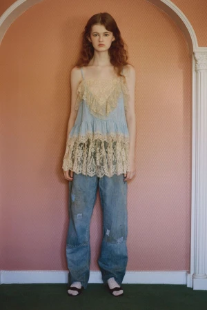 A young adult woman with long curly auburn hair poses in a studio wearing a lace-trimmed tank top and distressed denim jeans.