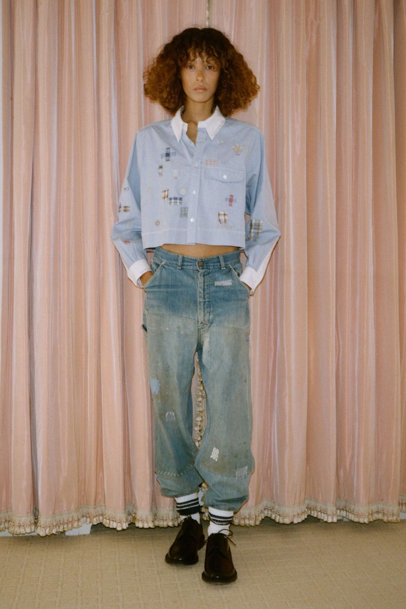 Full-length shot of an adult woman with curly hair, wearing a light blue collared shirt, distressed denim jeans, striped socks, and shoes.
