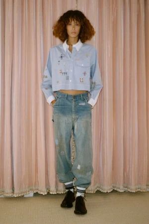 Full-length shot of an adult woman with curly hair, wearing a light blue collared shirt, distressed denim jeans, striped socks, and shoes.
