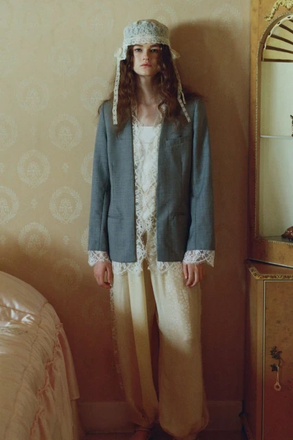 Adult female wearing a lace bonnet, a grey blazer, and loose fitting lace-trimmed yellow pants stands in an indoor setting.