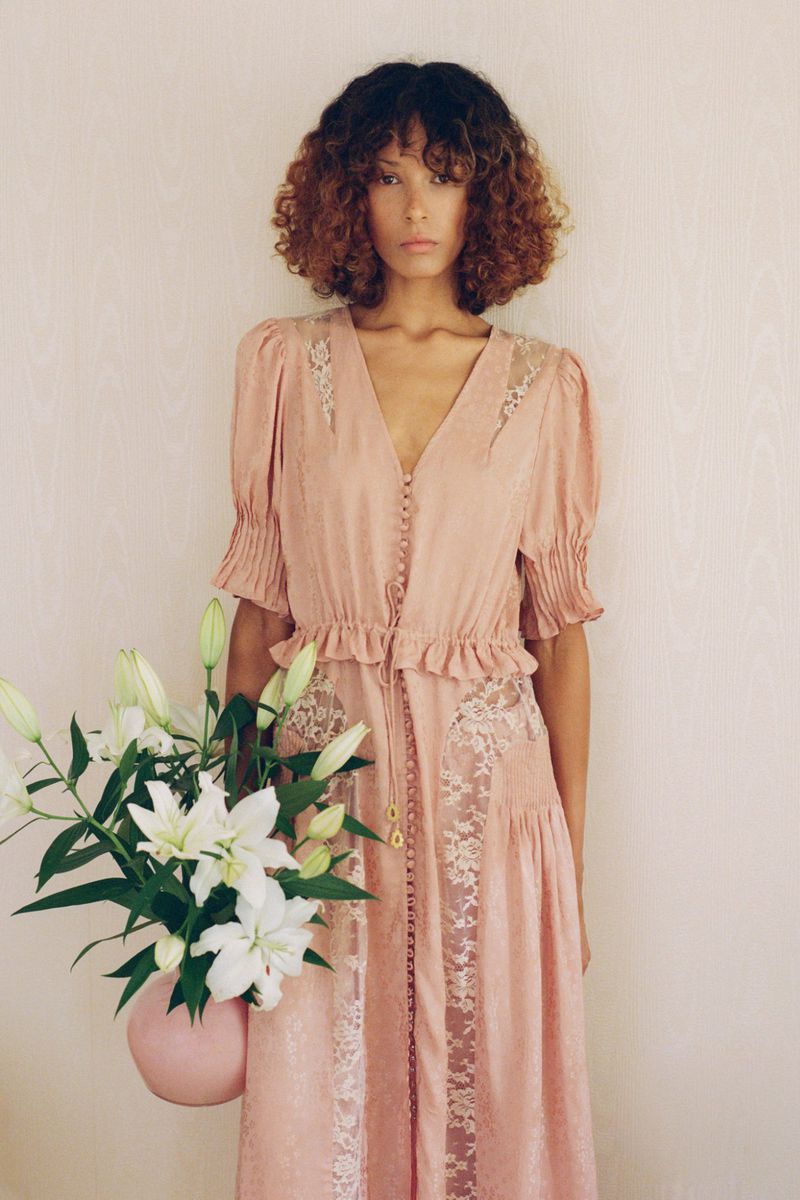 Woman in a pink gown poses with a vase of lilies in a studio setting, showcasing fashion and floral design with a clean aesthetic.