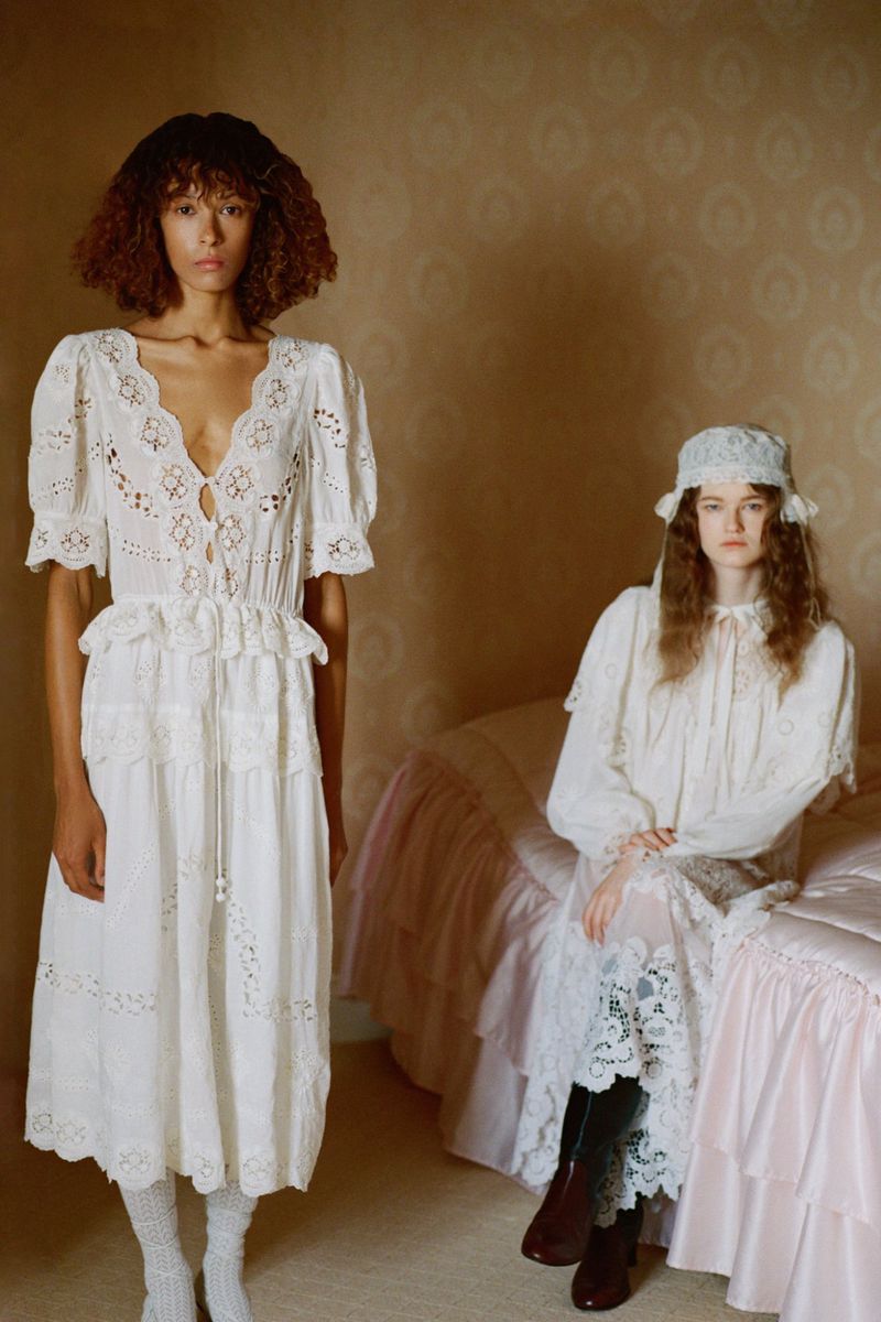 Two women pose indoors. One stands in a white eyelet dress and white socks; the other sits on a pink bed in a similar dress.
