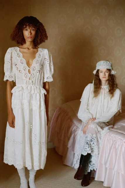 Two women pose indoors. One stands in a white eyelet dress and white socks; the other sits on a pink bed in a similar dress.