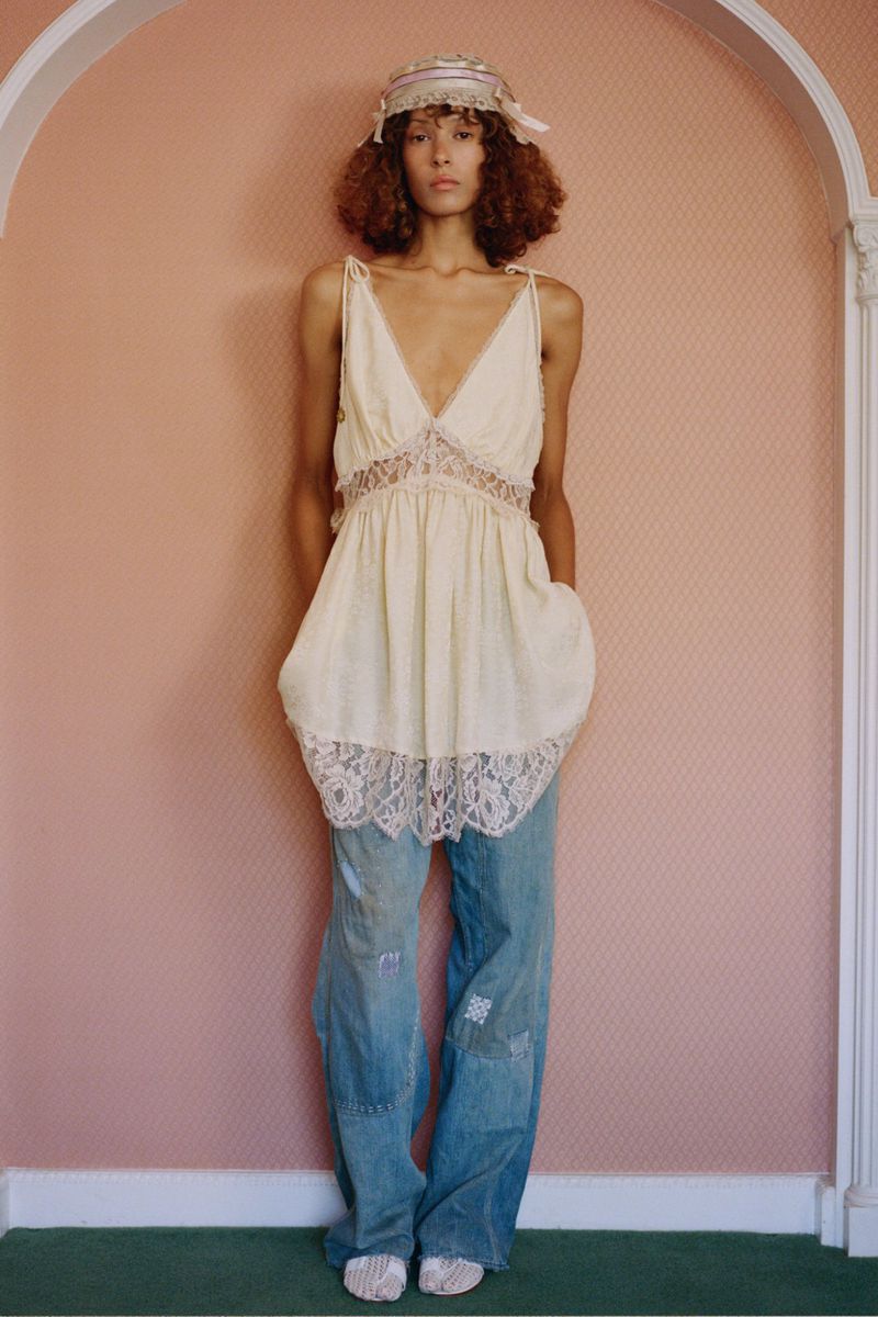 A curly-haired young woman poses in a cream lace-trimmed top and patchwork jeans against a pink wall, presenting a fashion-forward style.