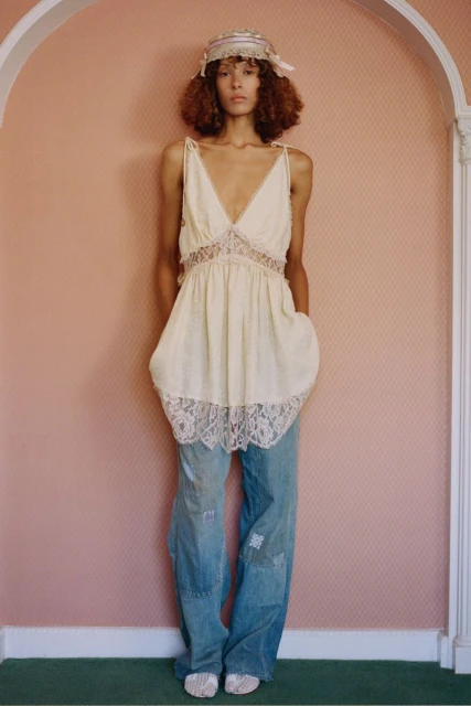 A curly-haired young woman poses in a cream lace-trimmed top and patchwork jeans against a pink wall, presenting a fashion-forward style.