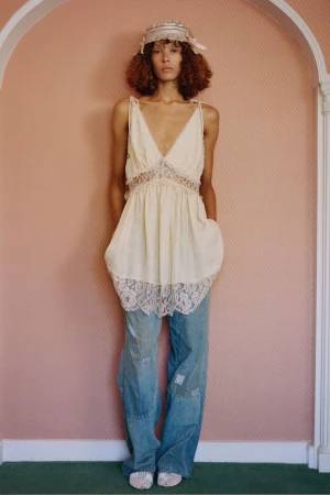 A curly-haired young woman poses in a cream lace-trimmed top and patchwork jeans against a pink wall, presenting a fashion-forward style.