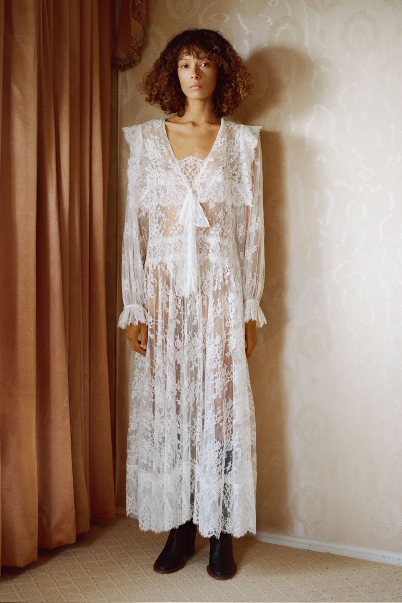Adult woman with curly hair wearing a white lace dress standing indoors near a curtain and wall, fashion advertisement.