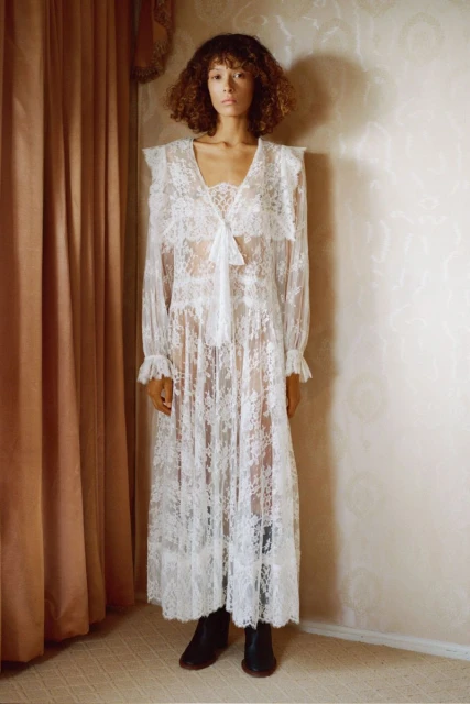 Adult woman with curly hair wearing a white lace dress standing indoors near a curtain and wall, fashion advertisement.