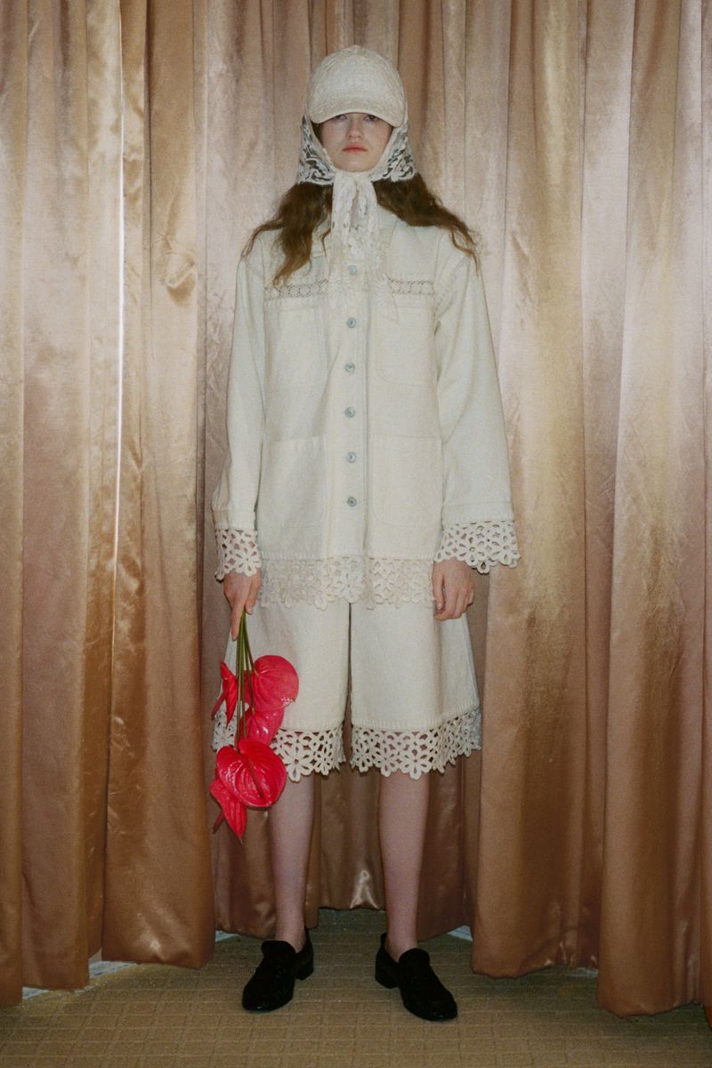 Woman poses indoors wearing white lace-trimmed outfit with a cap and holding red Anthurium flowers against a gold curtain backdrop.