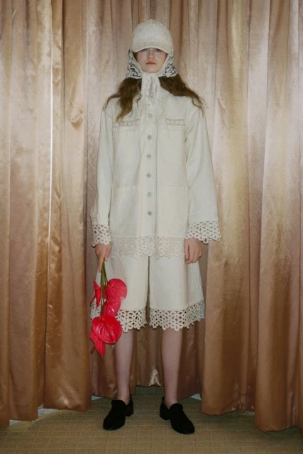 Woman poses indoors wearing white lace-trimmed outfit with a cap and holding red Anthurium flowers against a gold curtain backdrop.