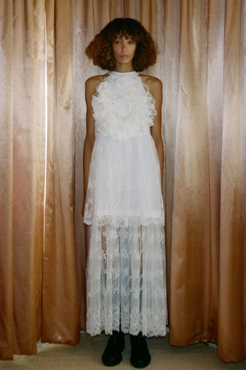 Adult woman wearing white, semi-sheer lace dress, with curly brown hair, posing against a neutral-toned curtain backdrop in a studio setting.
