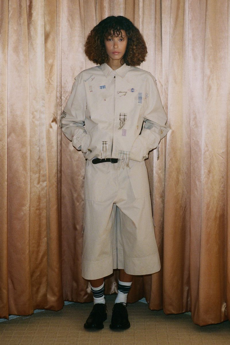 Full-shot of a model in a light beige shirt and culottes, standing against a draped curtain backdrop, showcasing fashion-forward style.