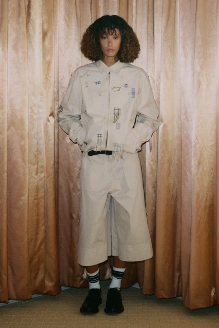 Full-shot of a model in a light beige shirt and culottes, standing against a draped curtain backdrop, showcasing fashion-forward style.