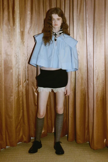 A fair-skinned young woman with auburn hair wears a blue top, black shorts, grey socks, and black shoes, posing indoors in a fashion shot.