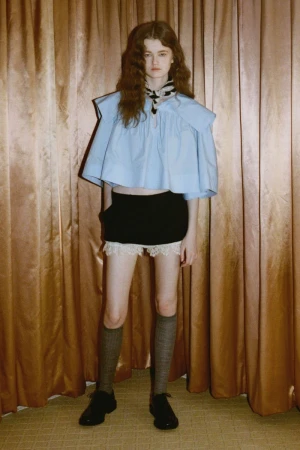 A fair-skinned young woman with auburn hair wears a blue top, black shorts, grey socks, and black shoes, posing indoors in a fashion shot.