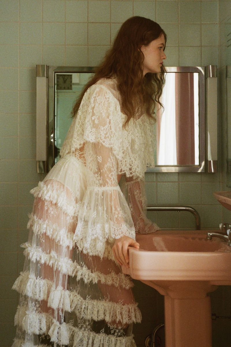 A young woman with auburn hair stands in a bathroom, wearing a tiered lace dress and leaning over a pink sink, looking away from the camera.