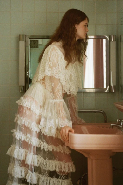 A young woman with auburn hair stands in a bathroom, wearing a tiered lace dress and leaning over a pink sink, looking away from the camera.