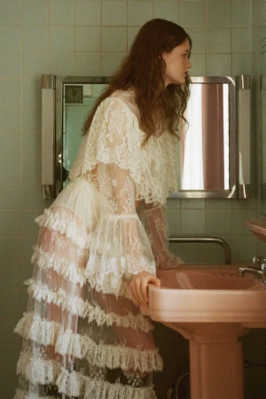 A young woman with auburn hair stands in a bathroom, wearing a tiered lace dress and leaning over a pink sink, looking away from the camera.