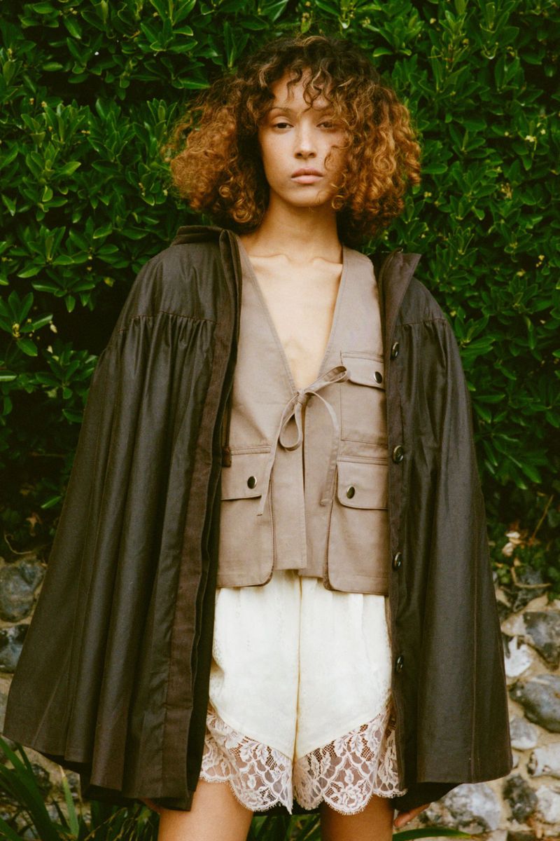 A young woman poses in a hooded cape, vest, and lace-trimmed shorts against a leafy green background in a fashion editorial shot.
