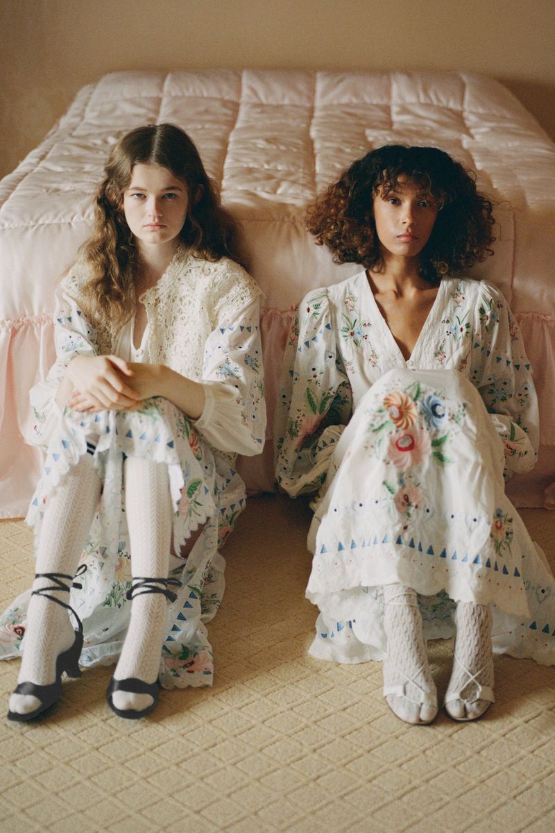 Two models seated on a carpeted floor in front of a quilted bed wearing long embroidered dresses and white socks.