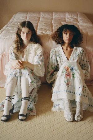 Two models seated on a carpeted floor in front of a quilted bed wearing long embroidered dresses and white socks.