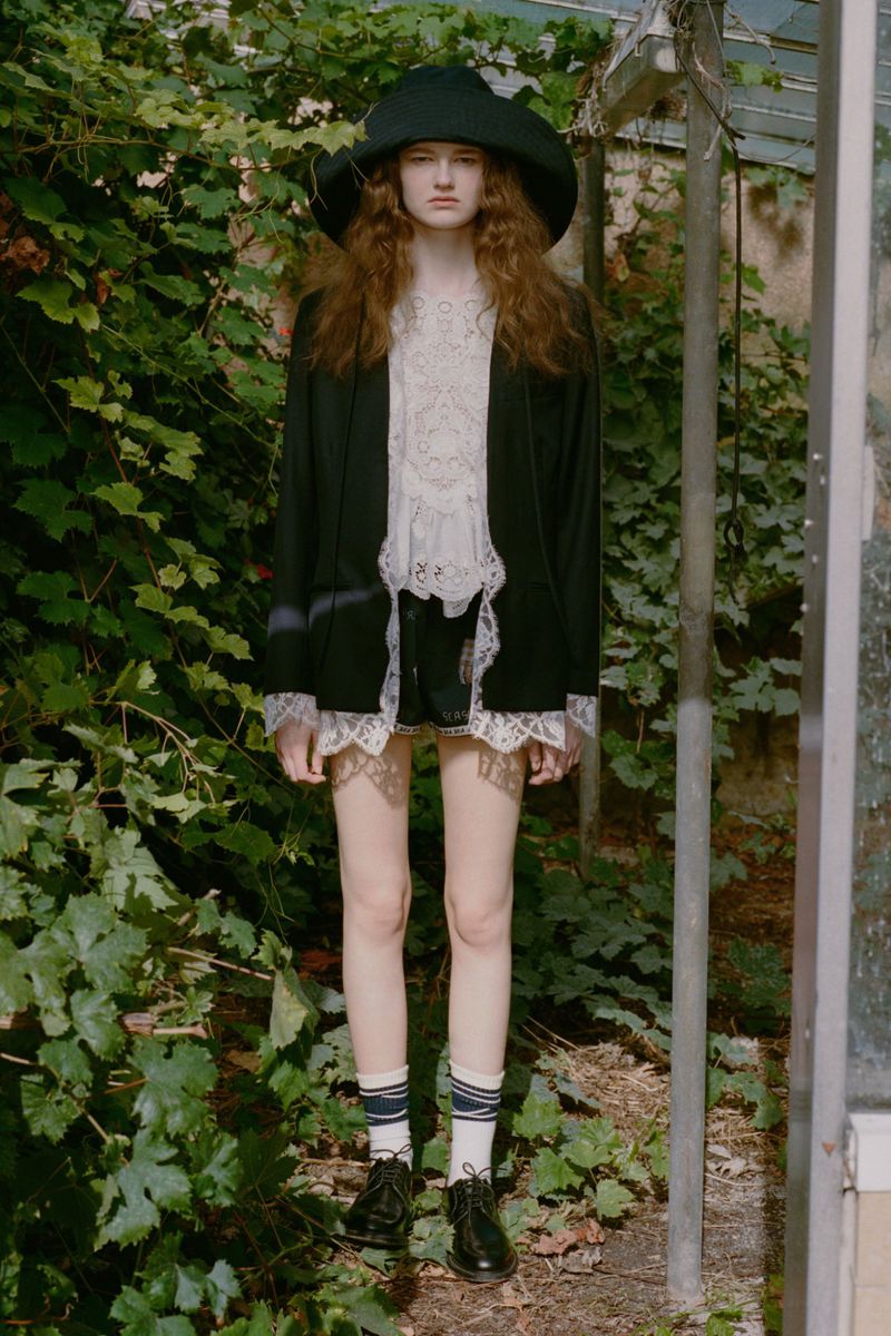 A young woman with wavy auburn hair, wearing a black jacket, shorts with lace trim, and a large dark hat, stands in a lush greenhouse.