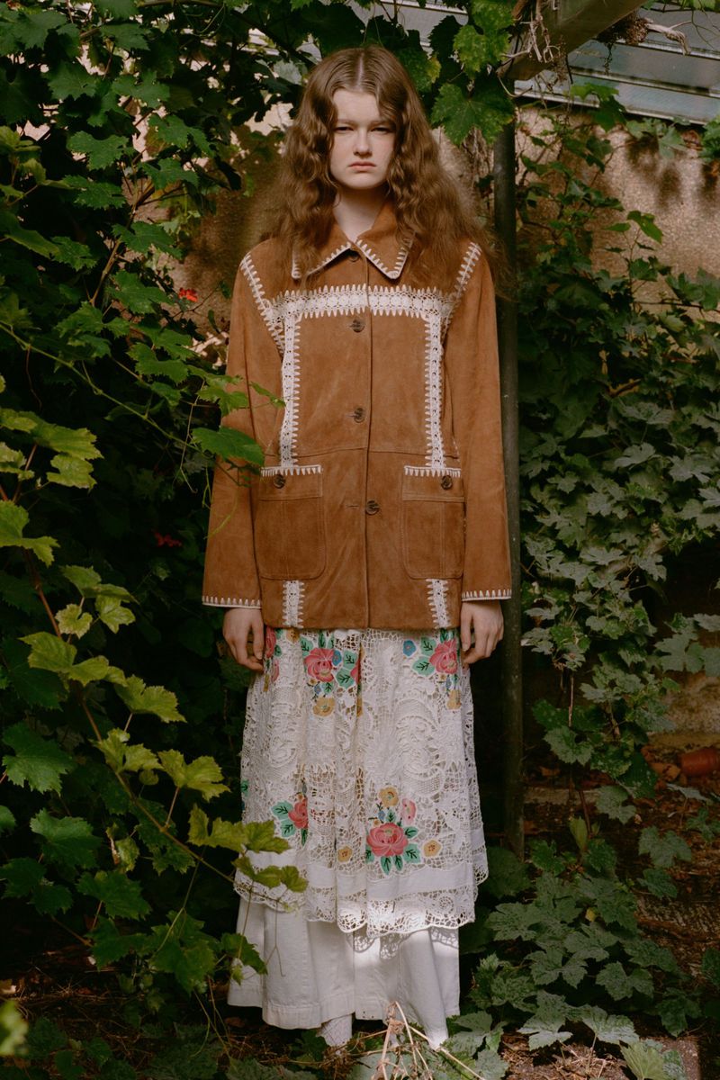 A young woman with long wavy hair poses outdoors, wearing a brown suede jacket with white embroidery and a lace floral skirt.