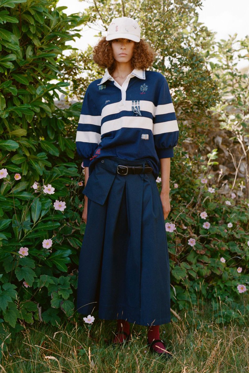 A young woman poses outdoors in a blue and white striped rugby shirt and navy skirt, complemented by a patterned hat and red socks.