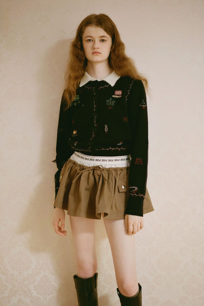 A young woman with long, wavy hair poses in a black sweater, khaki mini skirt, and black boots against a neutral patterned backdrop.