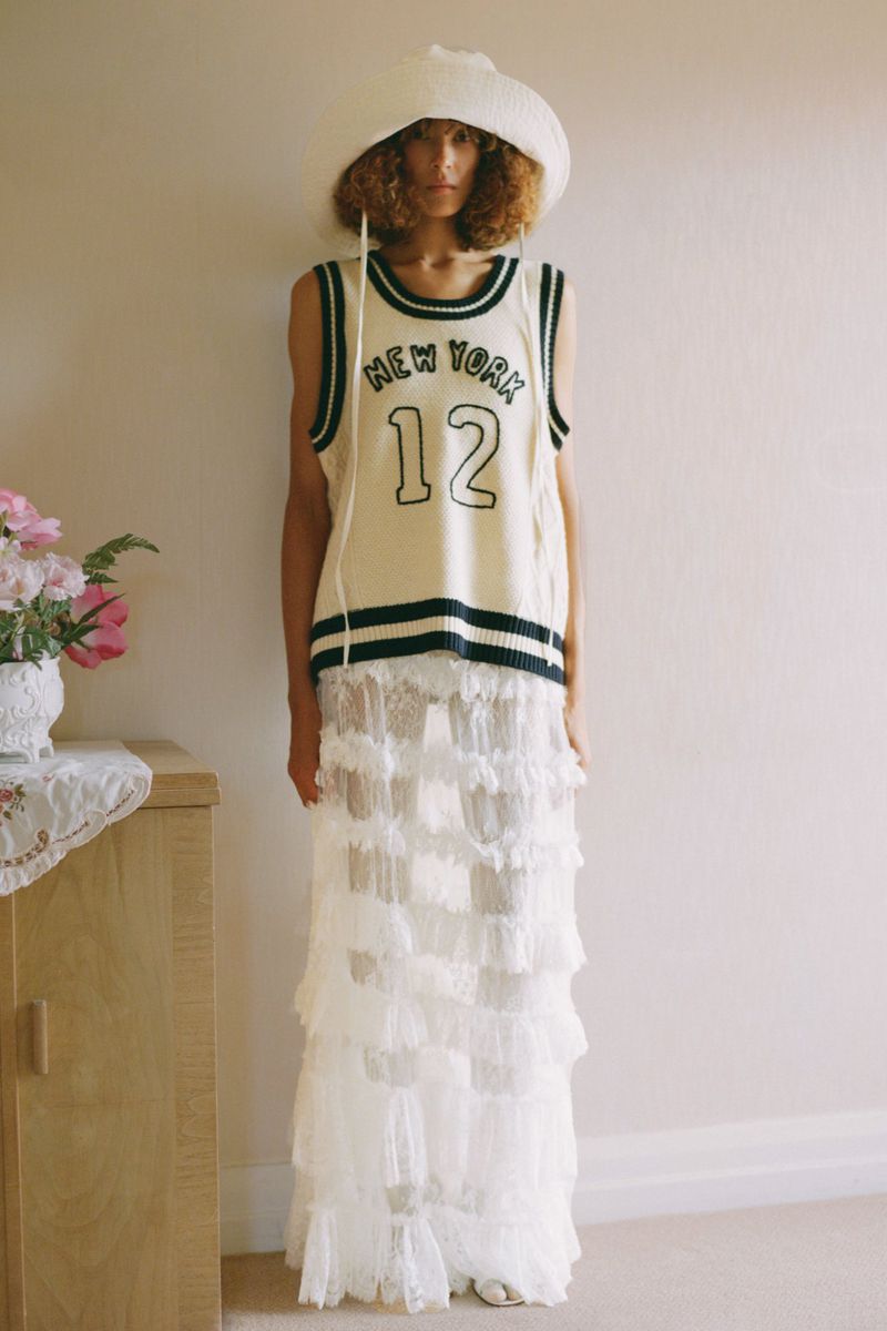 Adult female with curly hair wearing a white hat and knit 'New York 12' tank dress, posing in an indoor environment by a cabinet and flowers.