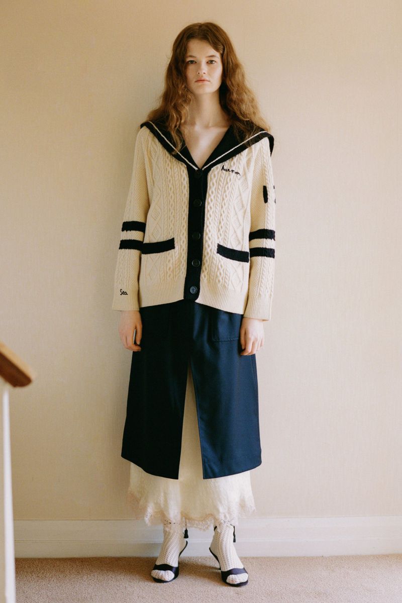 Full-length studio shot of a woman with curly hair wearing a cream cardigan, navy skirt, and white socks in front of a wall.