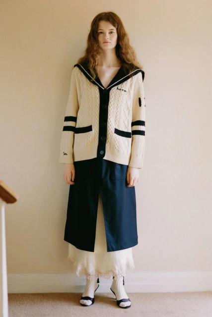Full-length studio shot of a woman with curly hair wearing a cream cardigan, navy skirt, and white socks in front of a wall.