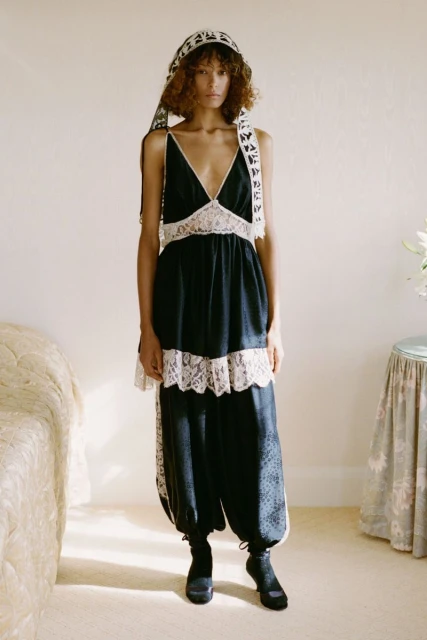 A woman poses in a black dress with lace trim and a matching headscarf indoors against a light wall with minimal props.