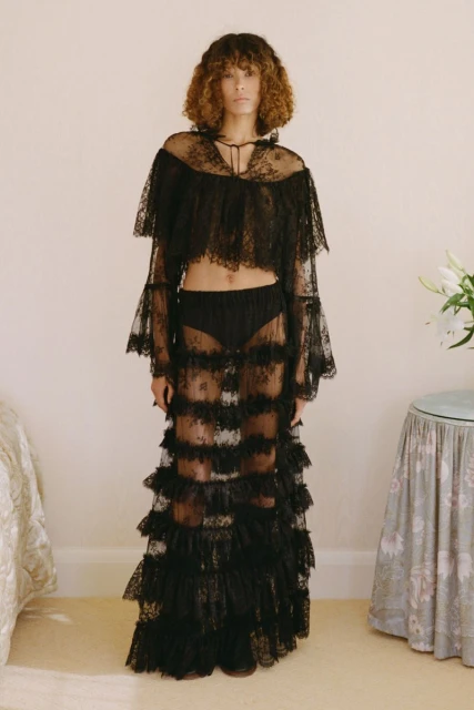 Fashionable adult woman poses in a sheer black lace top and skirt in an indoor setting with white walls and neutral carpeting.