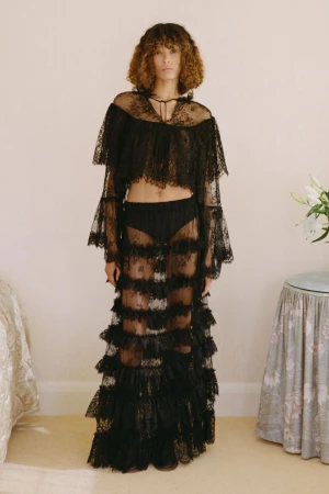 Fashionable adult woman poses in a sheer black lace top and skirt in an indoor setting with white walls and neutral carpeting.